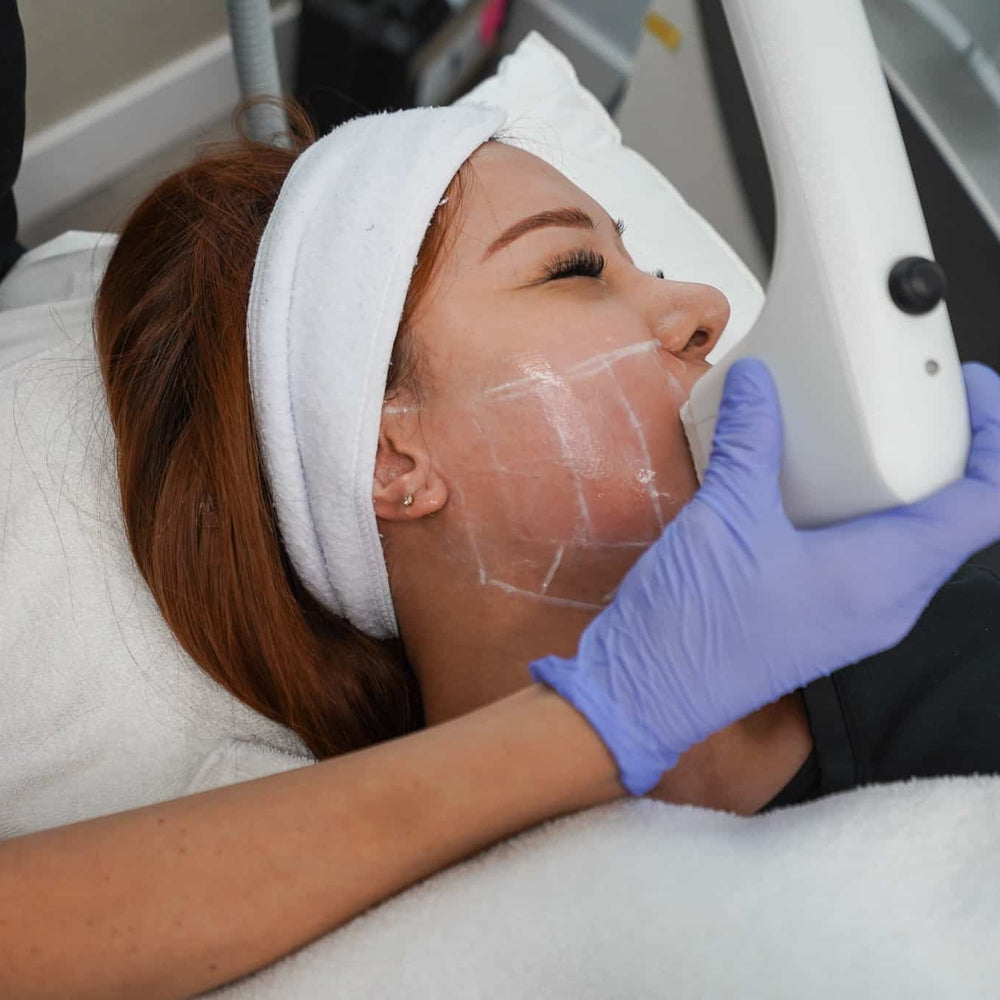 A person receiving a skin treatment, with a white headband, lying down, and a professional applying a device to the face.